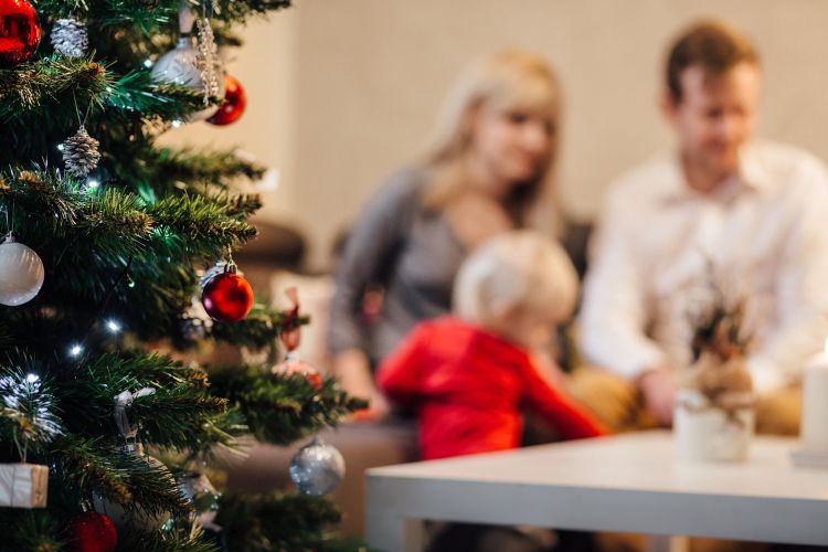 A Christmas tree with a mom and dad with a small toddler in the distance.
