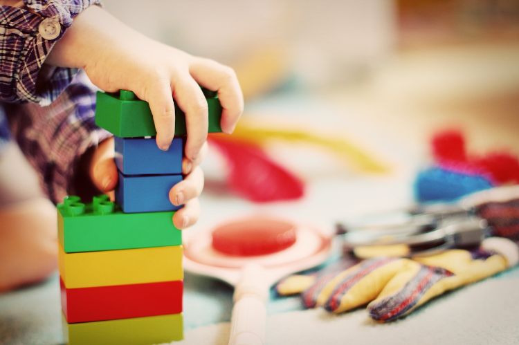 multiple colored legos stacked up with little kid hands holding them. Toys in the background.