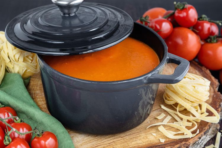 A photo of an orange colored soup in a cast iron pot placed on a cutting board surrounded by fresh tomatoes.
