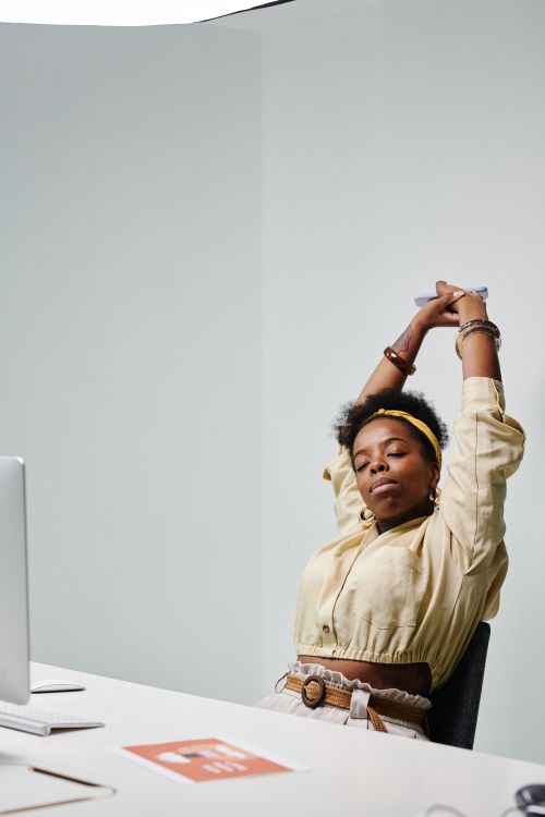 A woman stretches her arms up toward the ceiling while working at her desk.