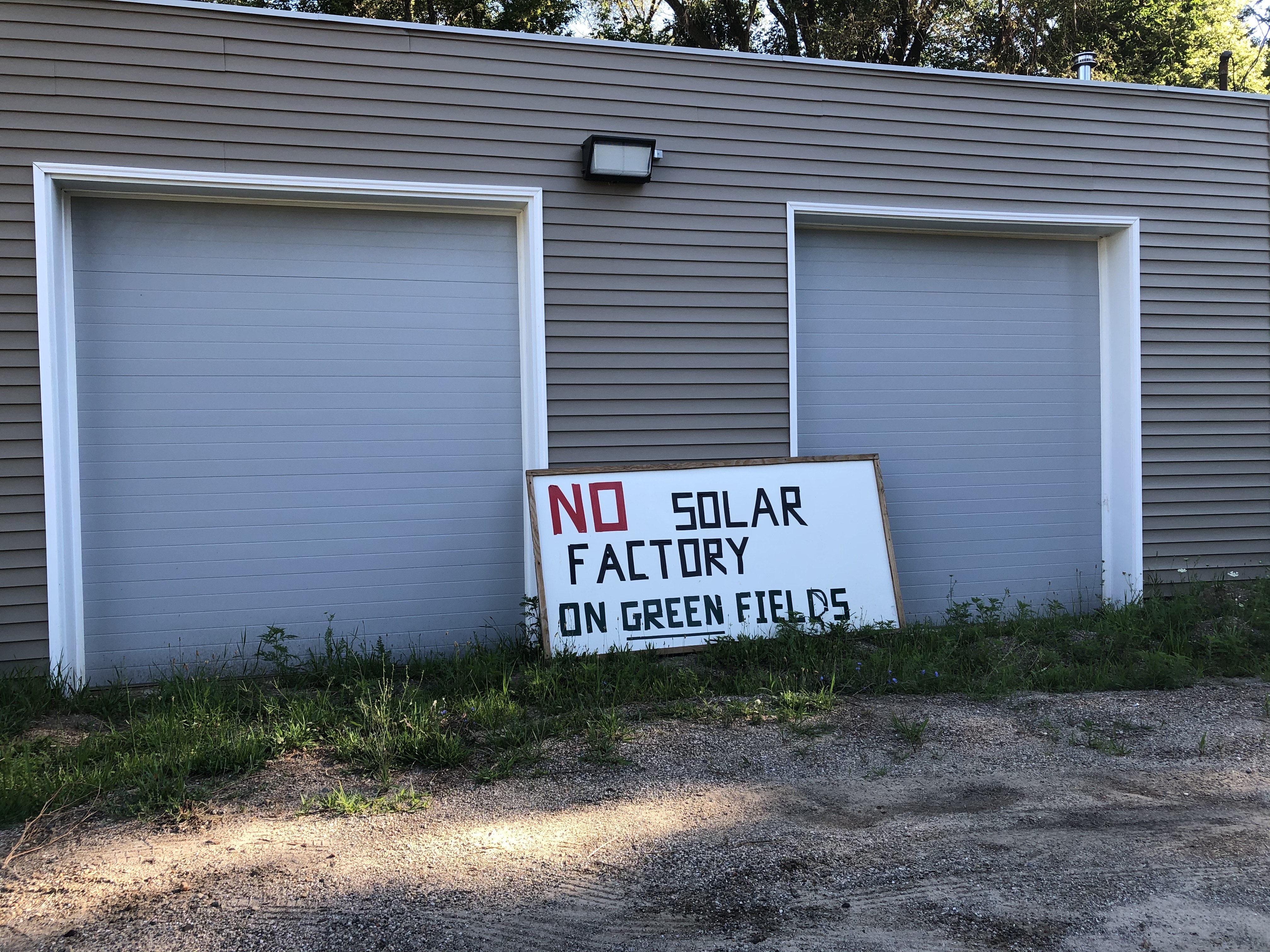Lettered yard sign saying &ldquo;No solar factory on green fields&rdquo; leaning against a beige building.