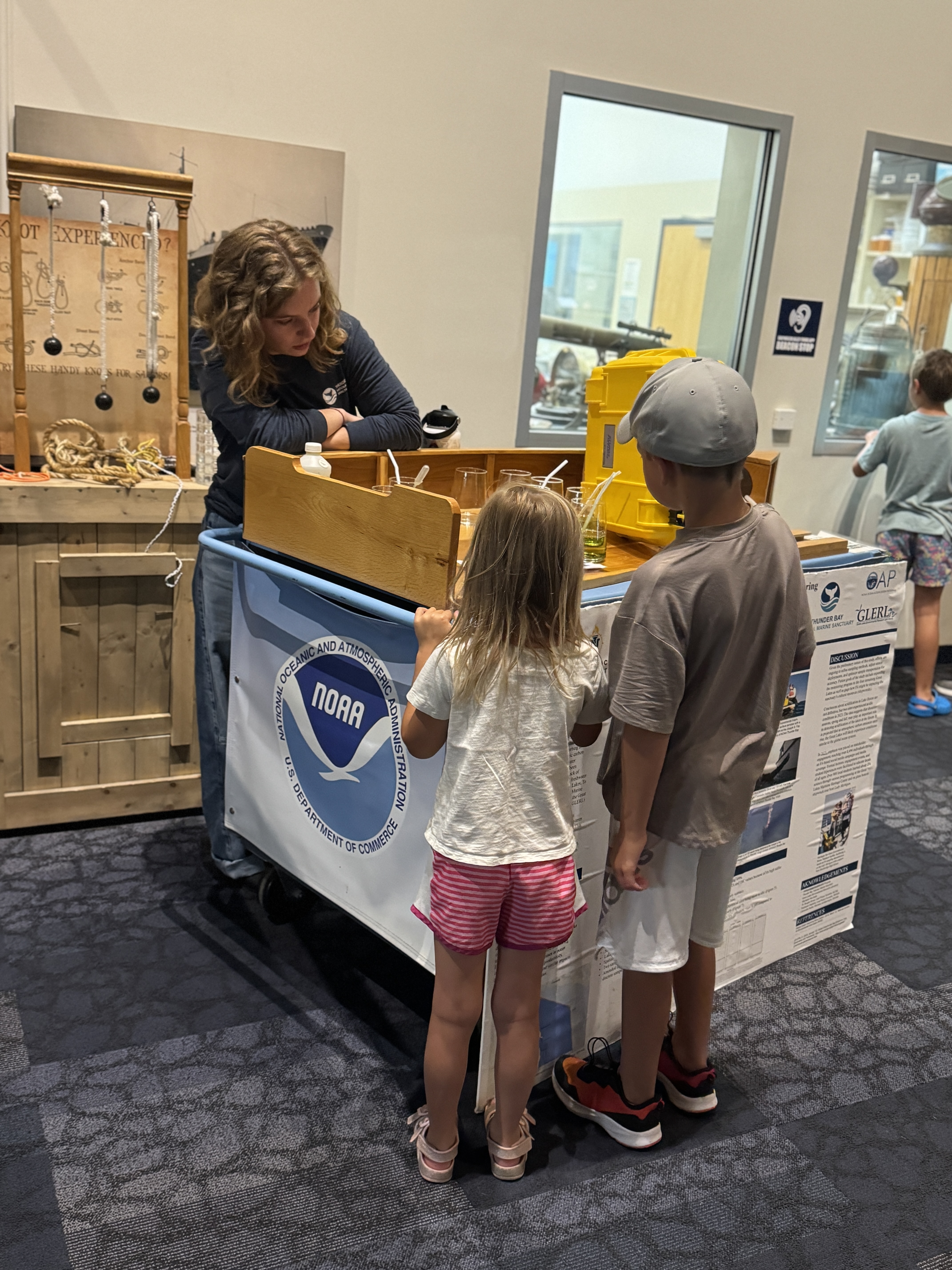 Woman stands at podium and shows several items to two children standing in front of the podium.